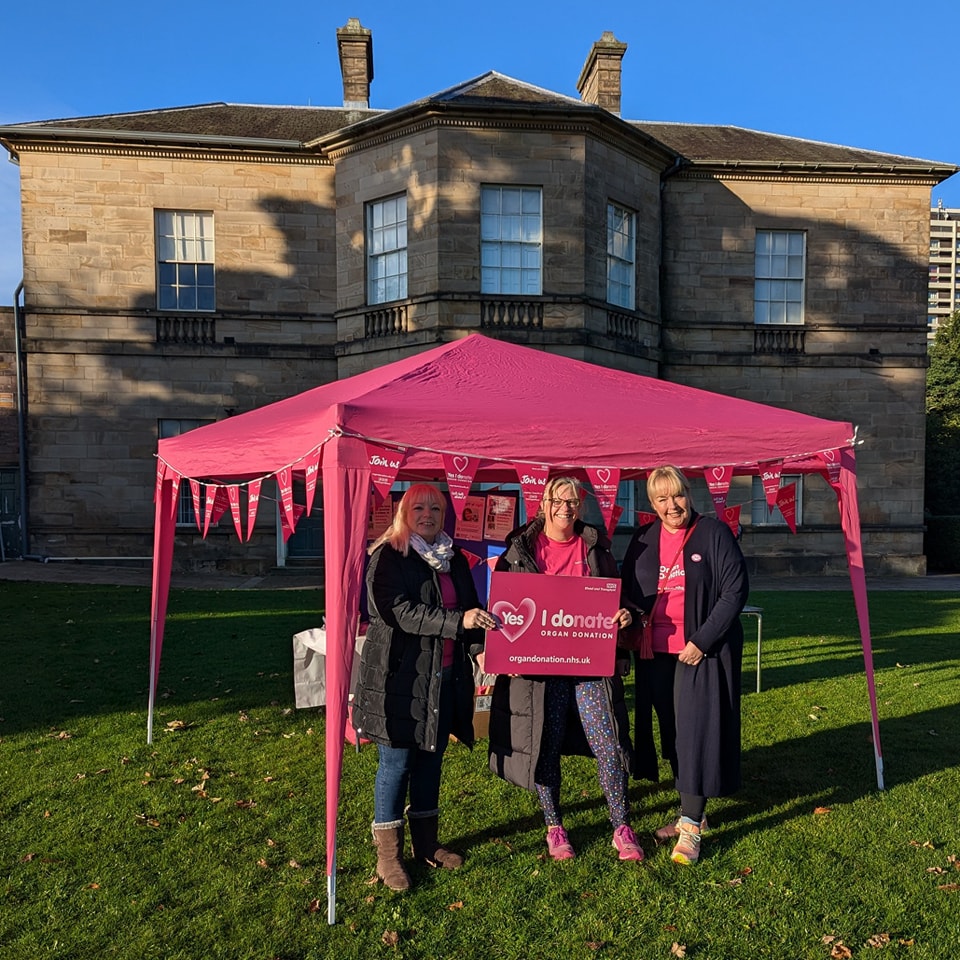 Three people holding a pink sign that says 'I donate' in front of a pink gazebo