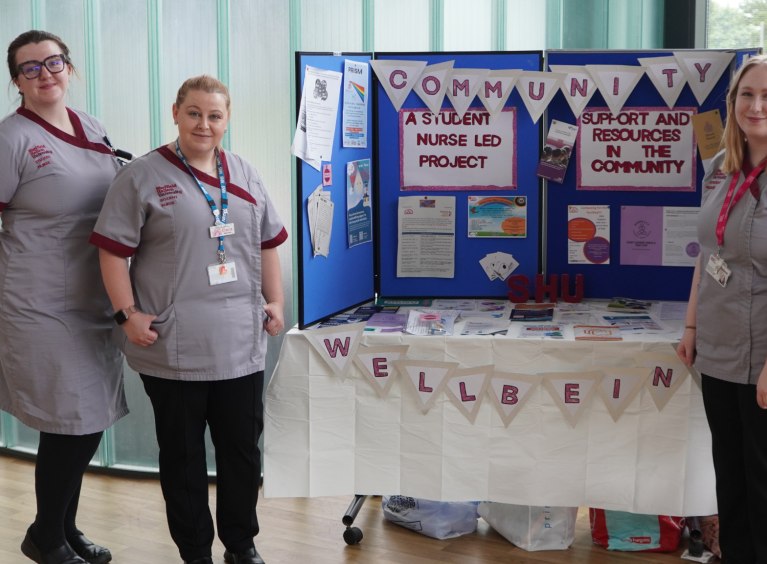 The image shows three student nurses standing by an information stand that promotes the services available to those experiencing loneliness.