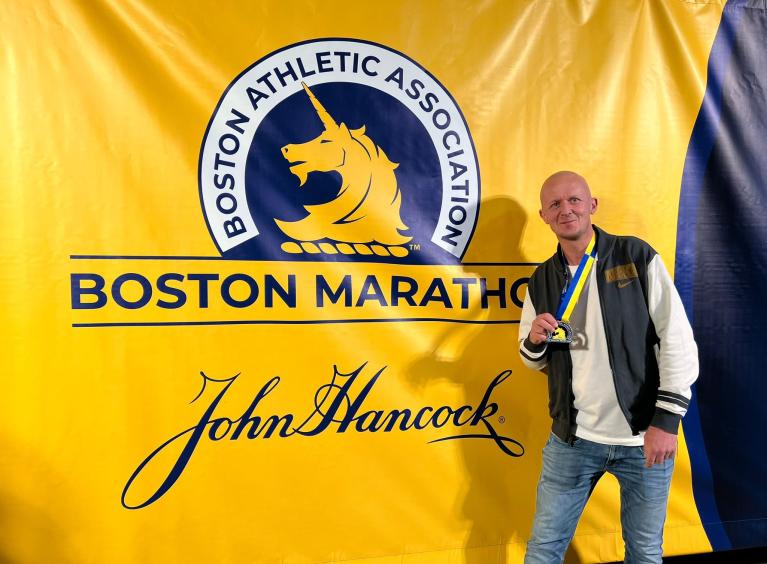 Steven Bryan is holding his Boston Marathon medal and is stood in front of a yellow sign with the Boston Marathon logo in blue