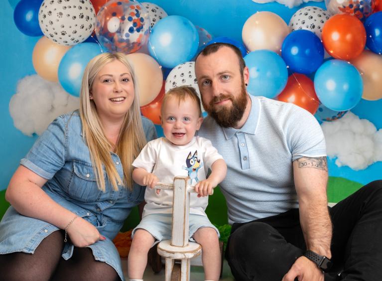 Teddy is sat between his parents. They are in front of balloons