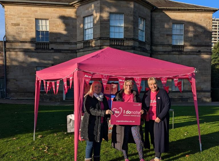 Three people holding a pink sign that says 'I donate' in front of a pink gazebo