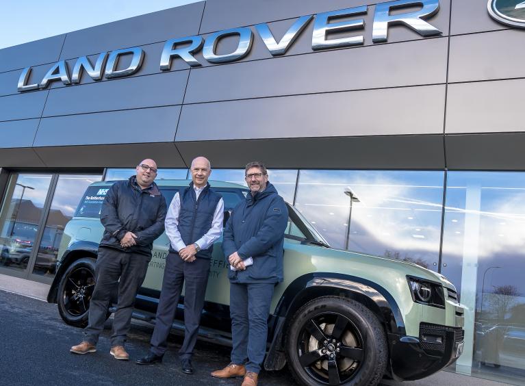 Three men stood in front of a light green Land Rover outside a showroom.