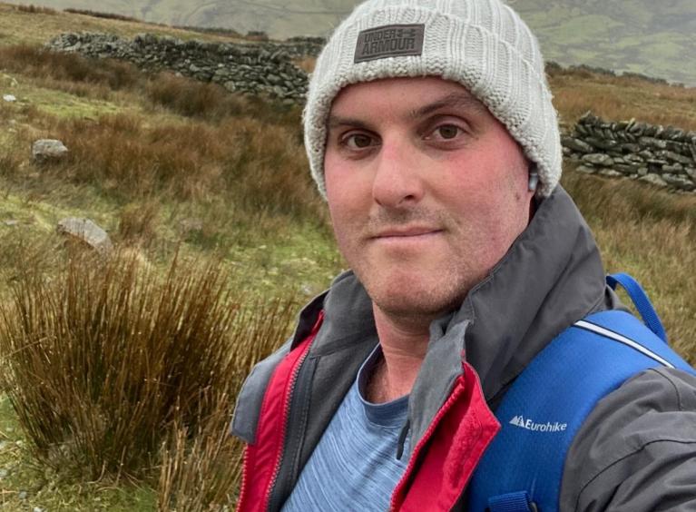 A man is wearing a coat, backpack and grey woollen hat. He is in the countryside with a peak behind him