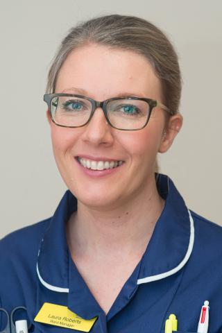 Laura is wearing a blue nursing uniform with a yellow name badge. She is wearing glasses with hair tied up.