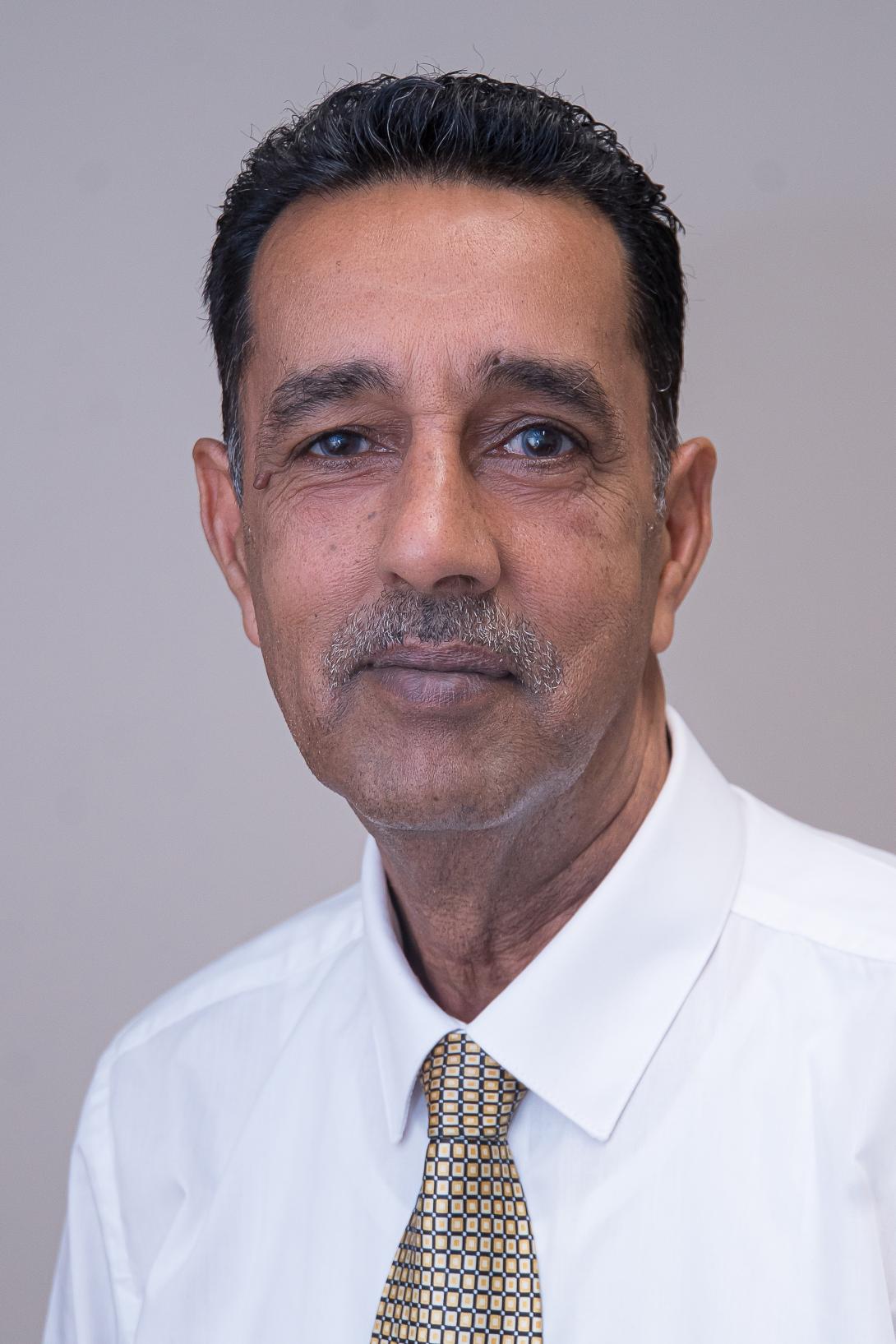 Mohammed is looking at the camera and is wearing a white shirt and brown tie with a squared pattern