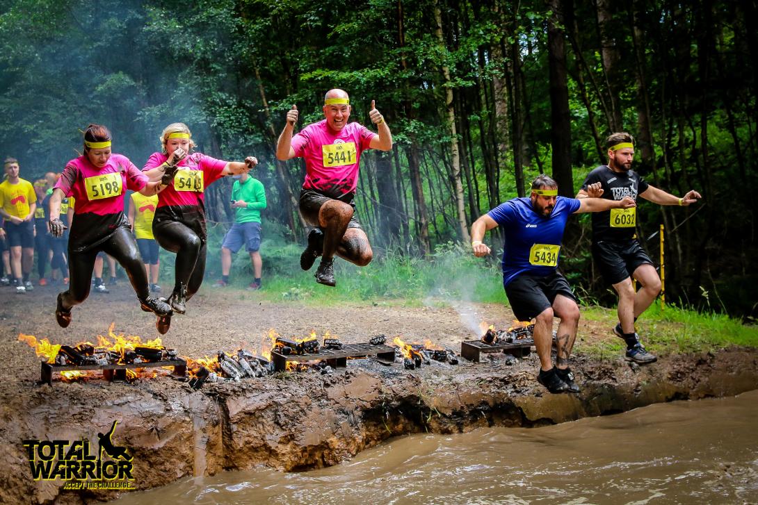 Five people, three in pink charity t-shirts, jump over fire into a muddy puddle