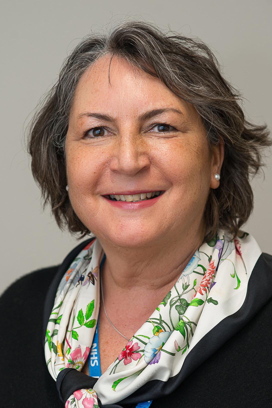 Angela is smiling at the camera. She has short hair and has a white scarf with a flower pattern around her neck