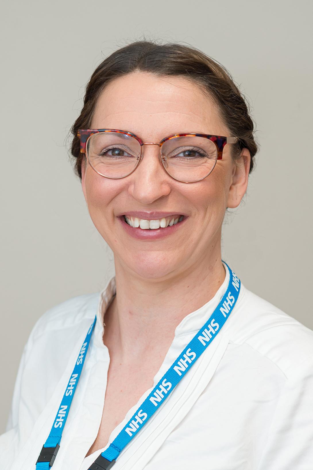 Sarah is smiling at the camera. She is wearing glasses, a white top and a blue NHS lanyard